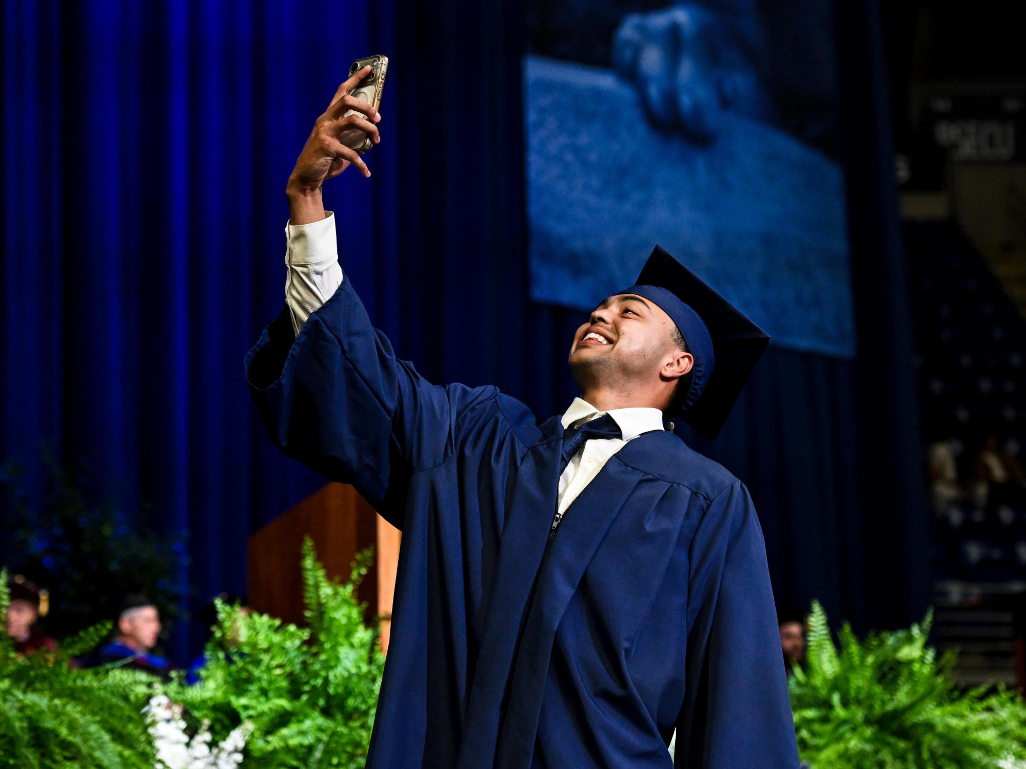 A student wearing a blue cap and gown takes a selfie