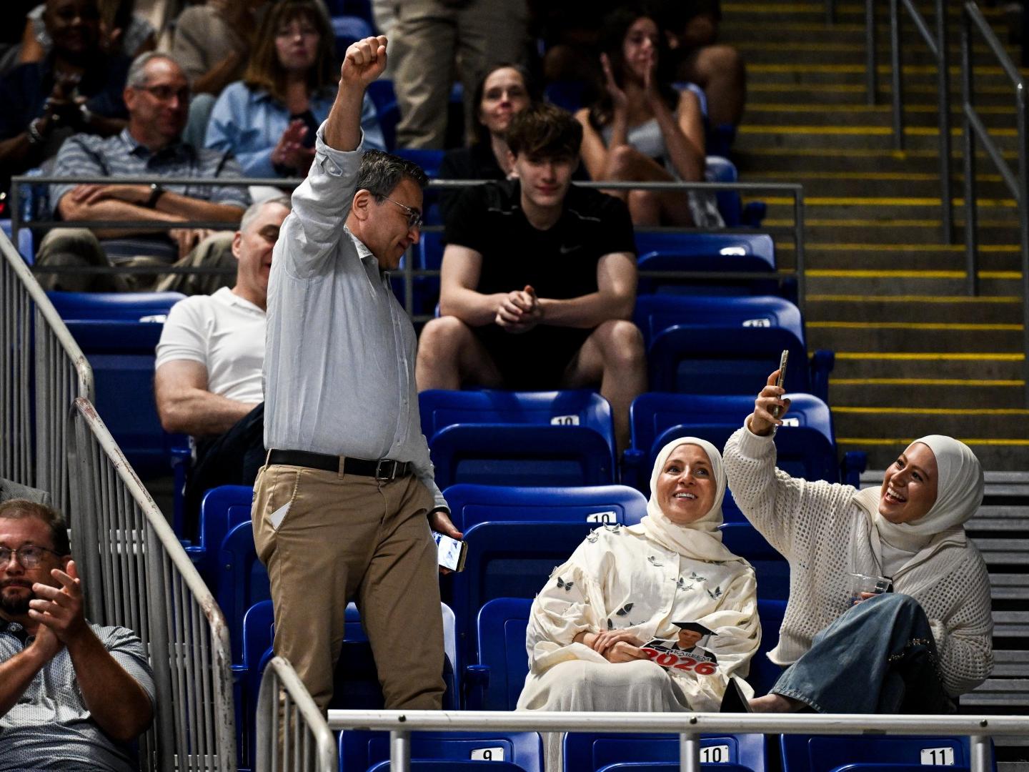 A man stands in celebration in an arena while a woman seated next to him takes his picture