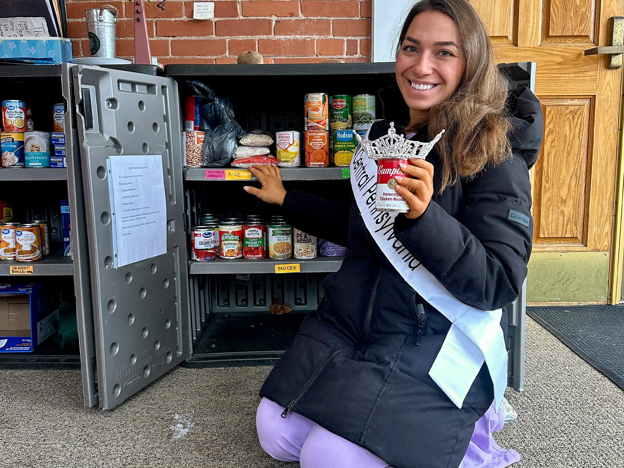 A woman wearing a crown and a sash puts canned goods in a pantry.