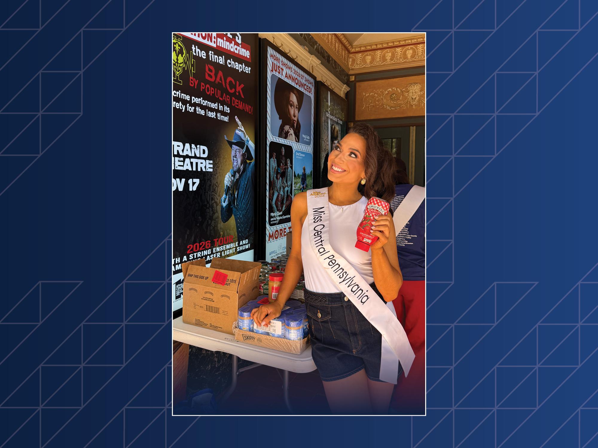 A woman wearing a sash and a crown poses next to a box of canned goods.