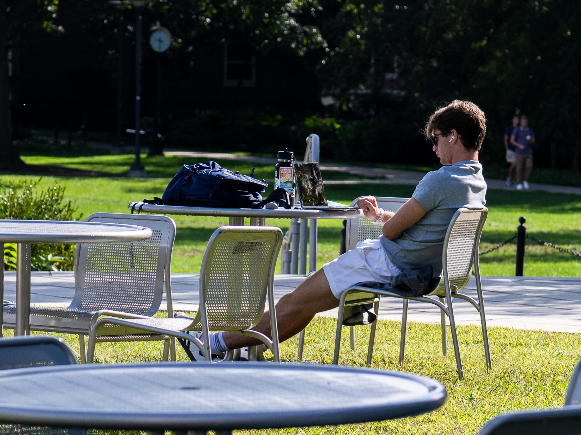 A student wearing sunglasses and earbuds sits at an outdoor table with a laptop and backpack on a sunny day at Penn State University Park