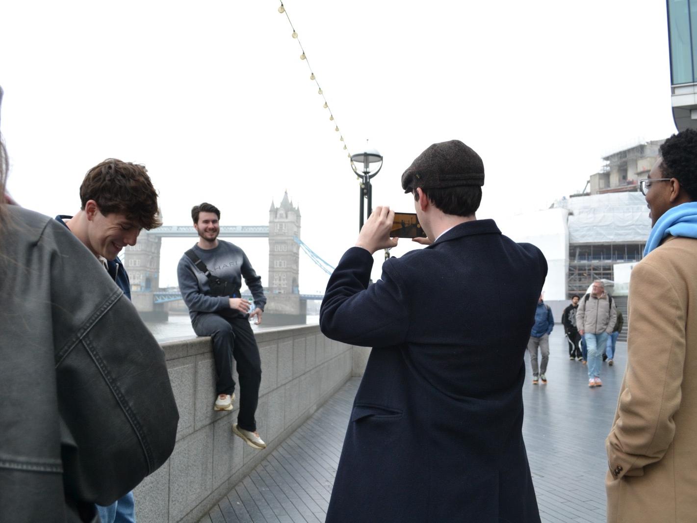 A male student in a jacket takes a picture of another male student who sits on a short wall
