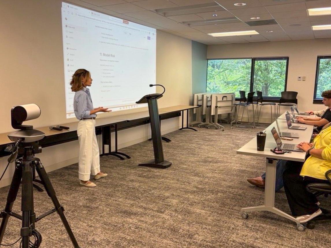 Emine Barik presents at a podium in a classroom, with a large screen displaying "Model Risk." Attendees sit at desks with laptops while a camera on a tripod captures the presentation.
