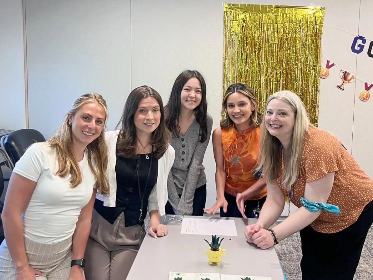 Five women smile and pose around a table in an office, with a yellow pineapple planter and decorative gold fringe in the background.