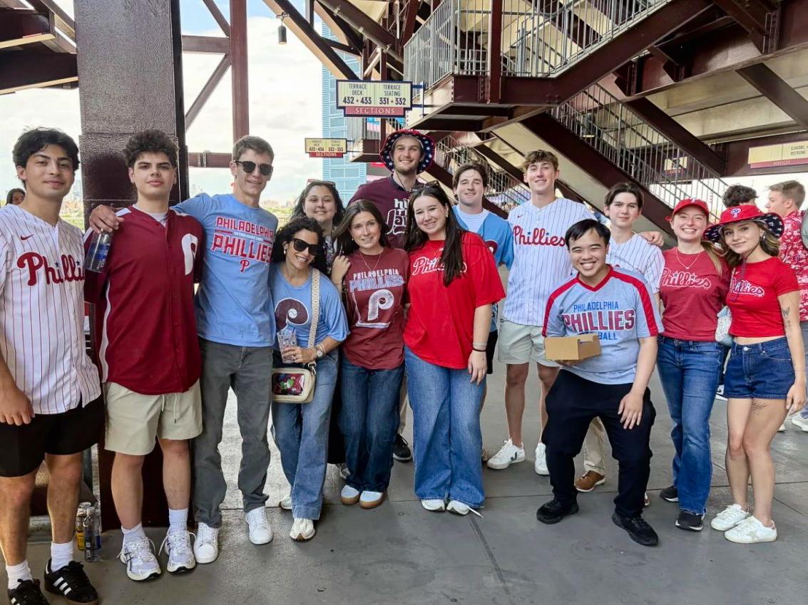 A group of young people wearing Philadelphia Phillies shirts and jerseys poses together at a stadium.