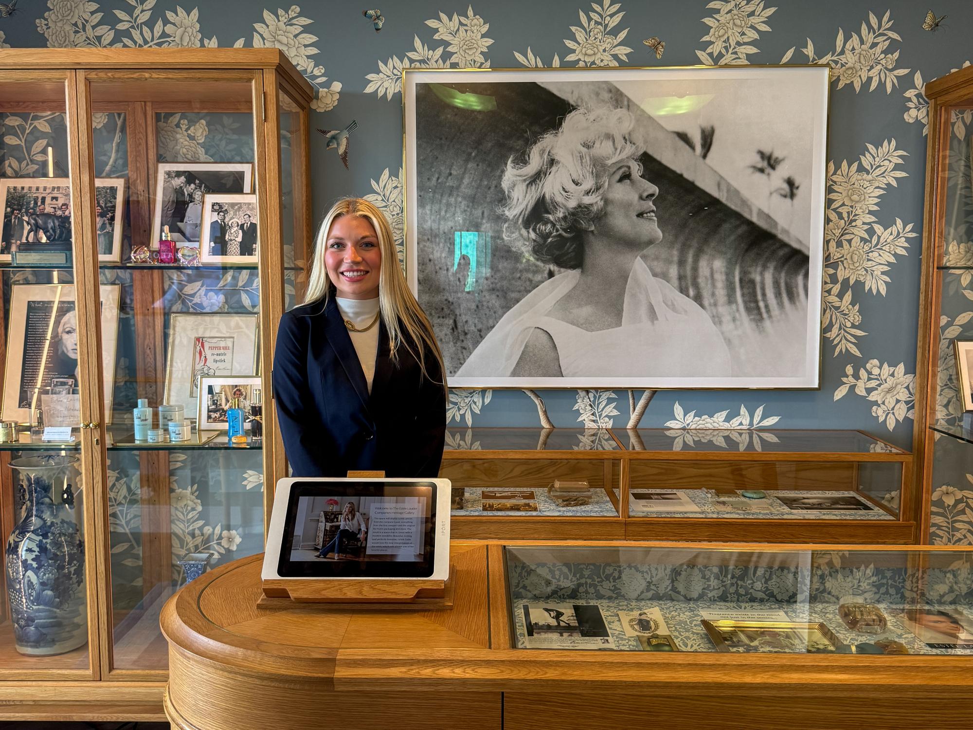Karli Brenneman stands at a wooden display desk in front of a large black-and-white portrait of Estée Lauder, surrounded by glass cabinets of vintage memorabilia.