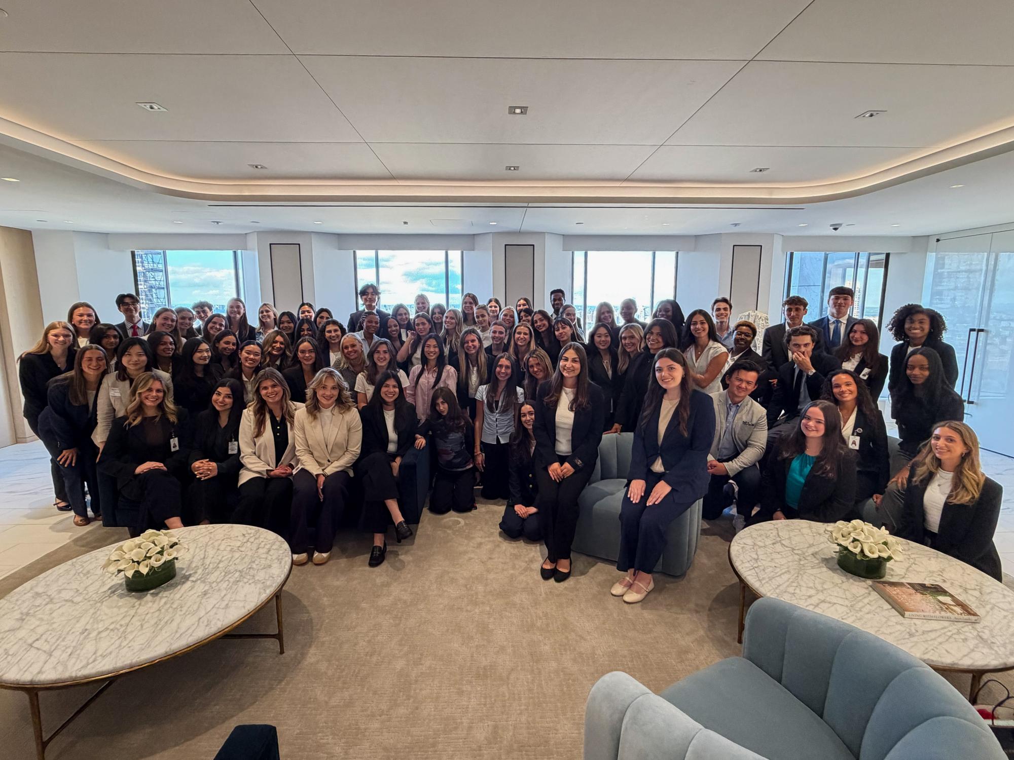 A large group of interns and staff pose together in a corporate lounge with floor-to-ceiling windows, seated and standing around marble coffee tables.