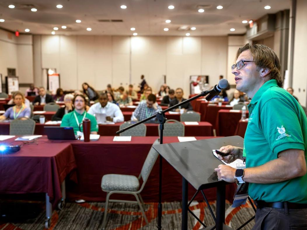 A man stands at a podium during a presentation he's giving to a large event space filled with people sitting at rows of tables.