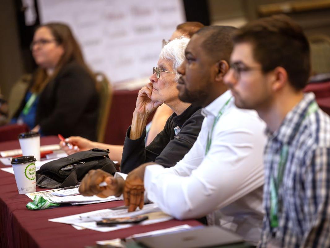 A close-up image of a row of people seated at a table, listening to a presentation.