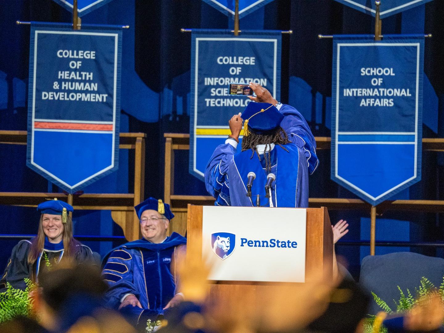 A person wearing a blue cap and gown takes a selfie with his back toward the photographer
