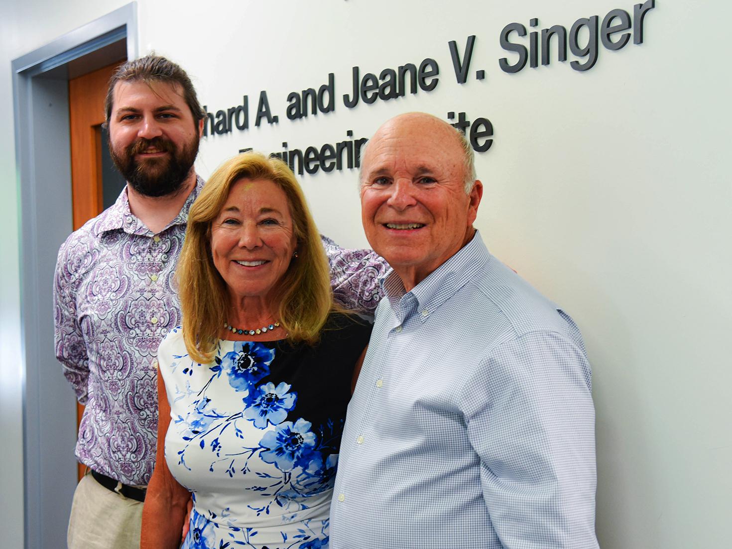 Scott Singer, Jeane Singer, and Richard Singer smiling in front of a hallway plaque.