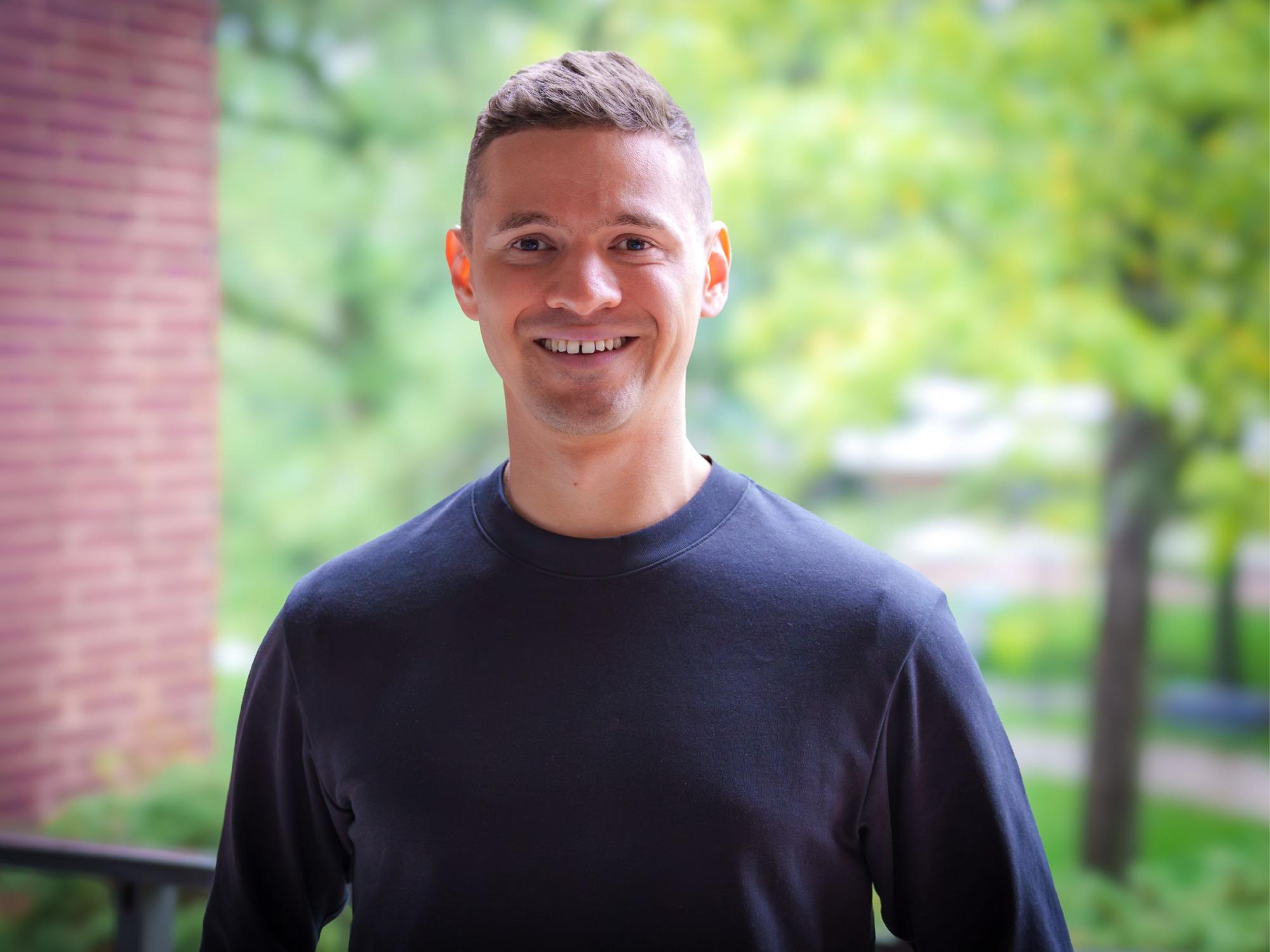 smiling person with short dark hair and navy blue shirt with red brick wall and green trees in background