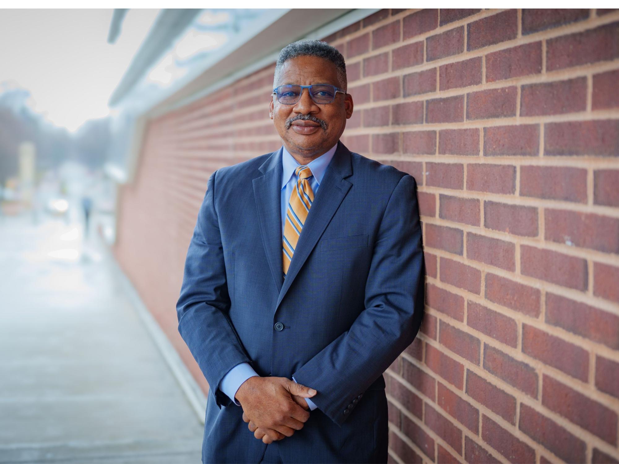 college professor in business attire leans against red brick wall