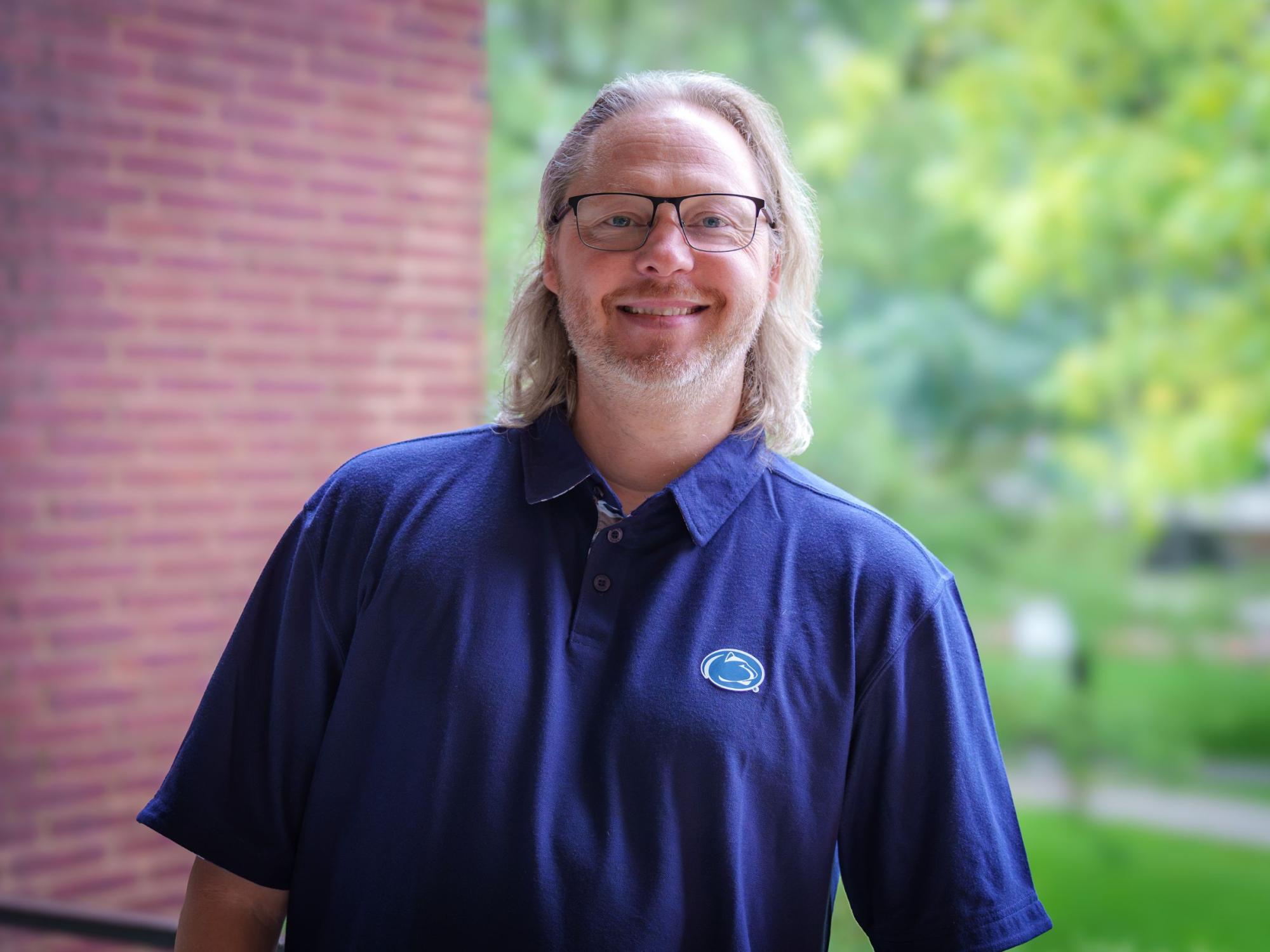 person with long gray hair, gray goatee, wire-frame glasses and navy blue Penn State golf shirt