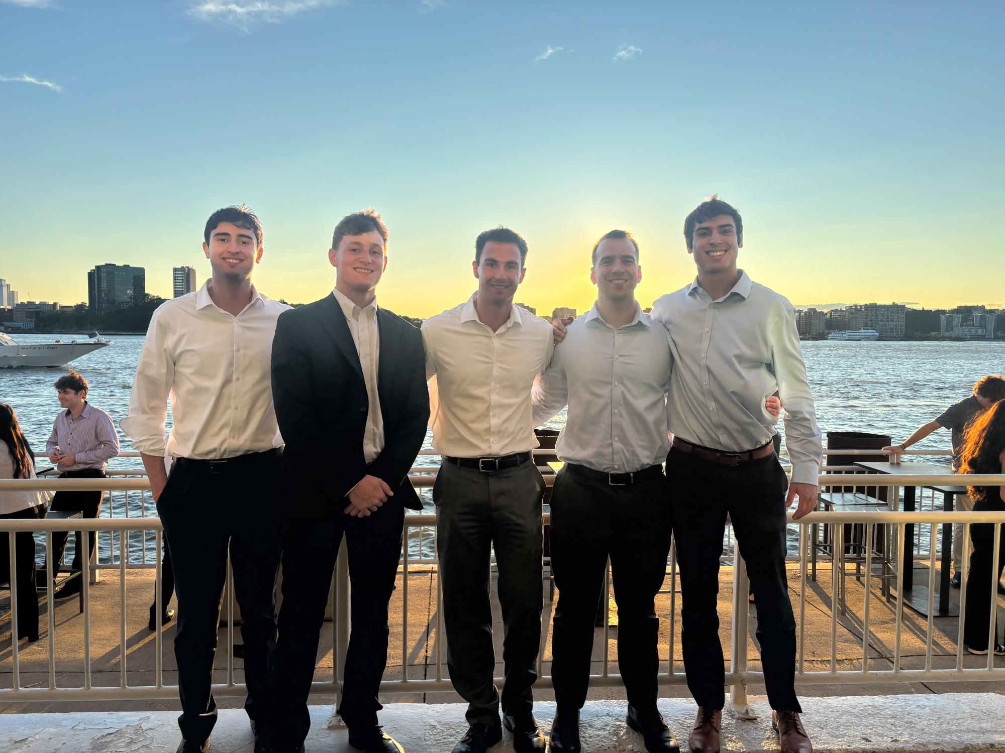 Five young men in business attire stand in front of a railing with water and a city skyline behind them.