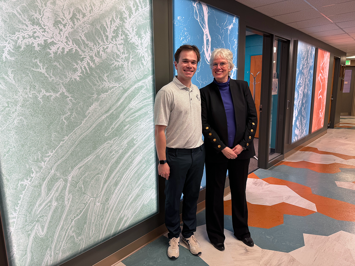 Nate and Cindy stand smiling in front of a green map wall panel in the Walker building, with a colorful geometric floor design.