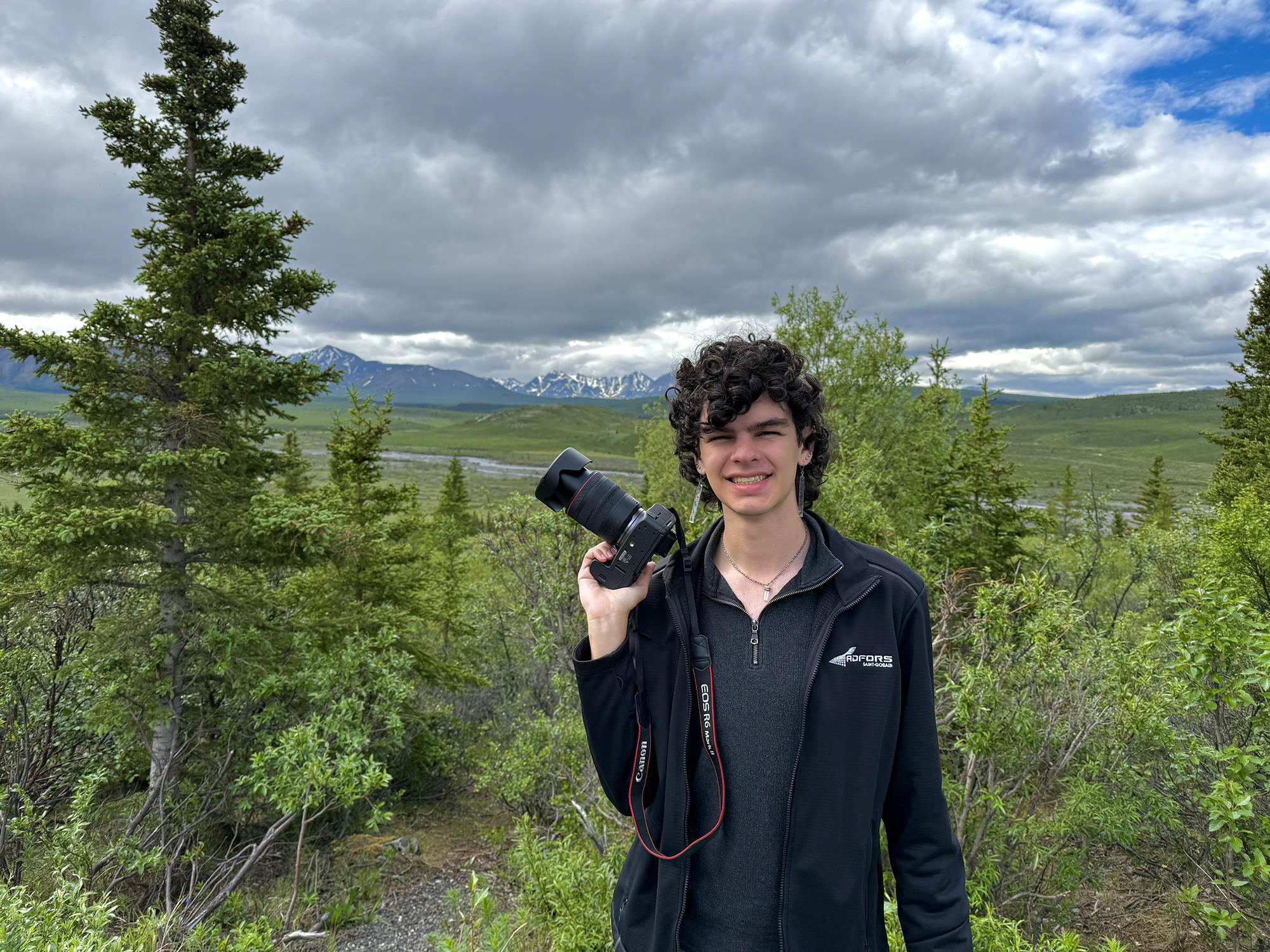 Student with a camera walks Alaskan landscape with mountains in view overhead.
