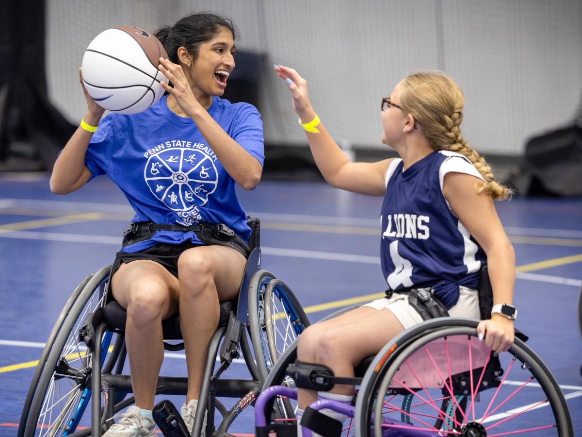 An individual in a wheelchair holds a basketball in the air and smiles. Another person to her right reaches out toward the ball.