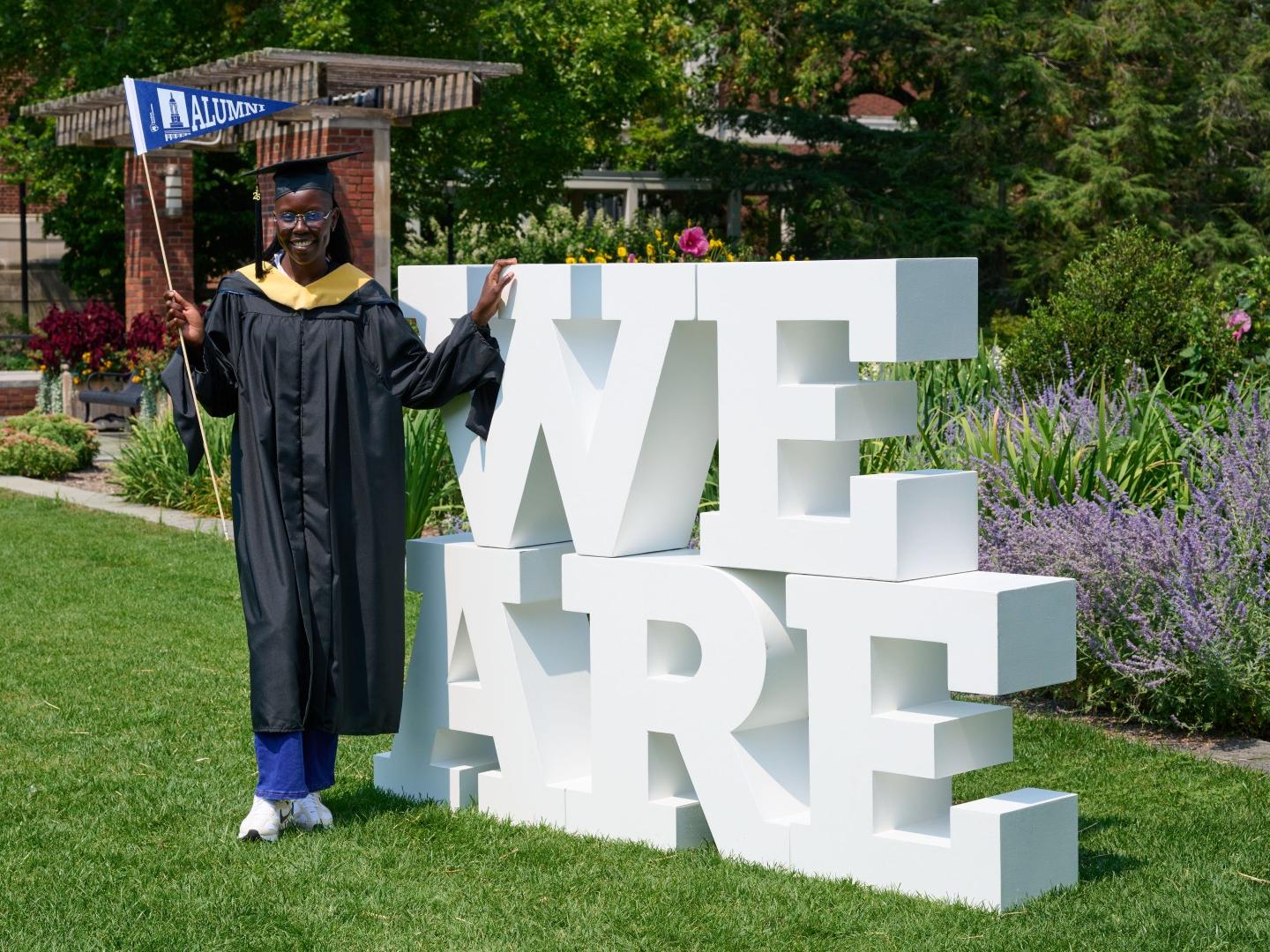 A student wearing a black cap and gown holds an "Alumni" pennant while standing next to a large "We Are" sign