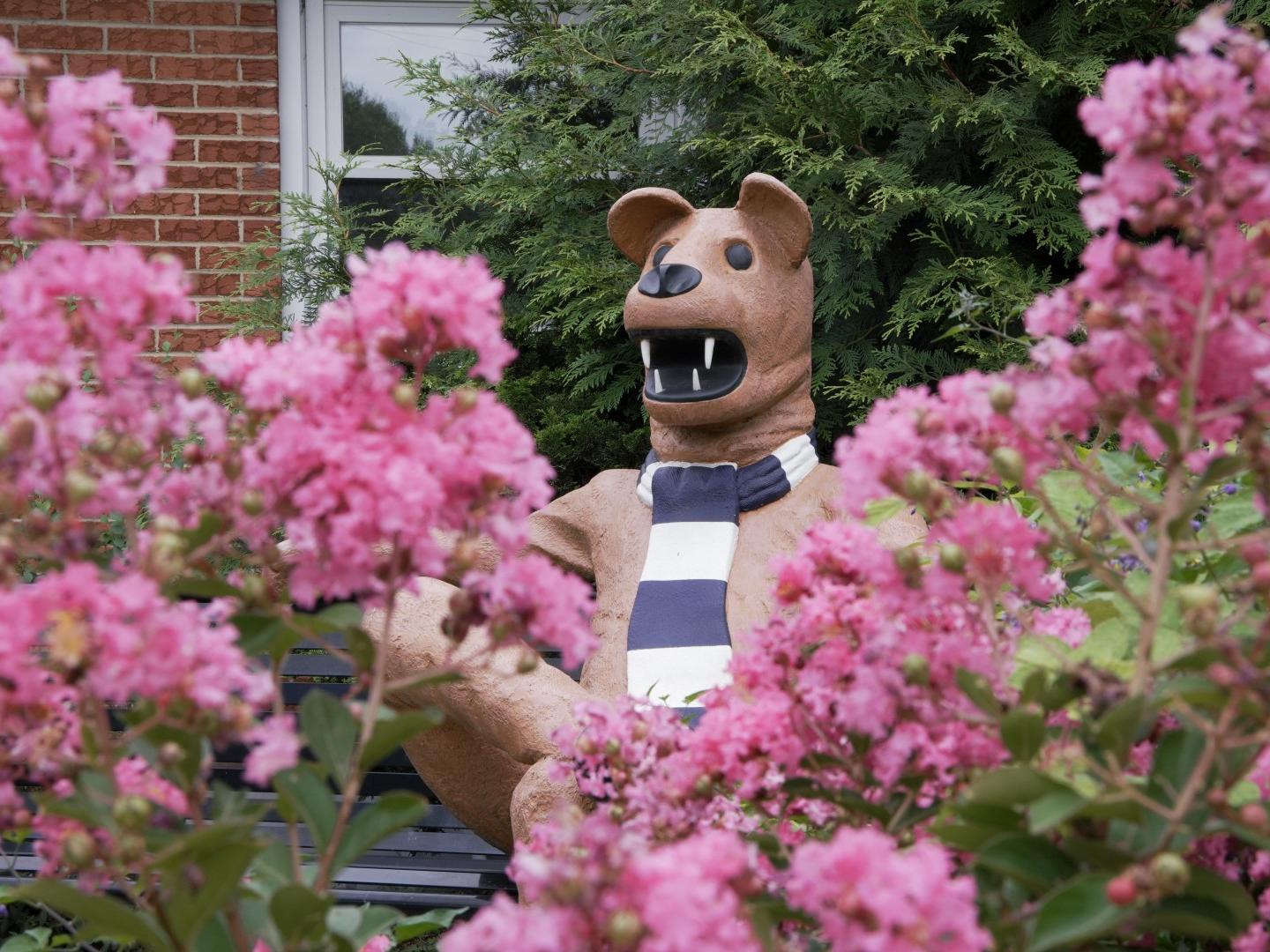 A Nittany Lion statue sits on a bench between pink flowers