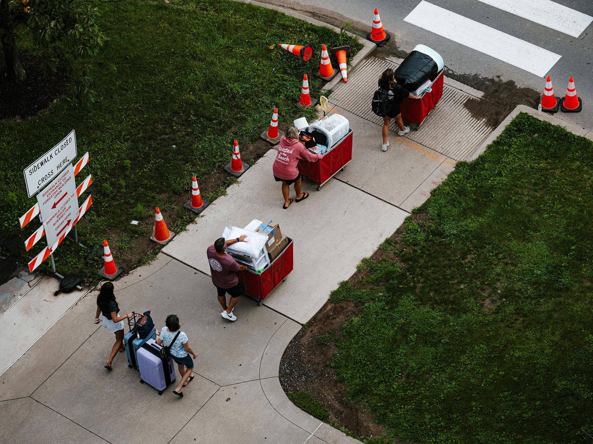 Four students pushing large carts full of household goods approach a crosswalk lined with traffic cones