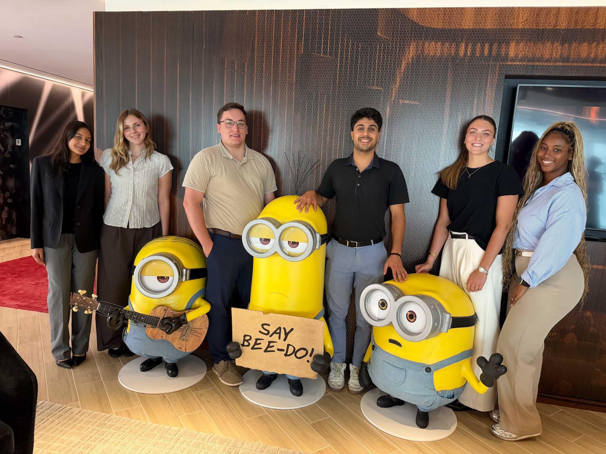 Reid-Pitta stands second from the left wearing a white top and brown pants. She is standing in front of Minion statues. 