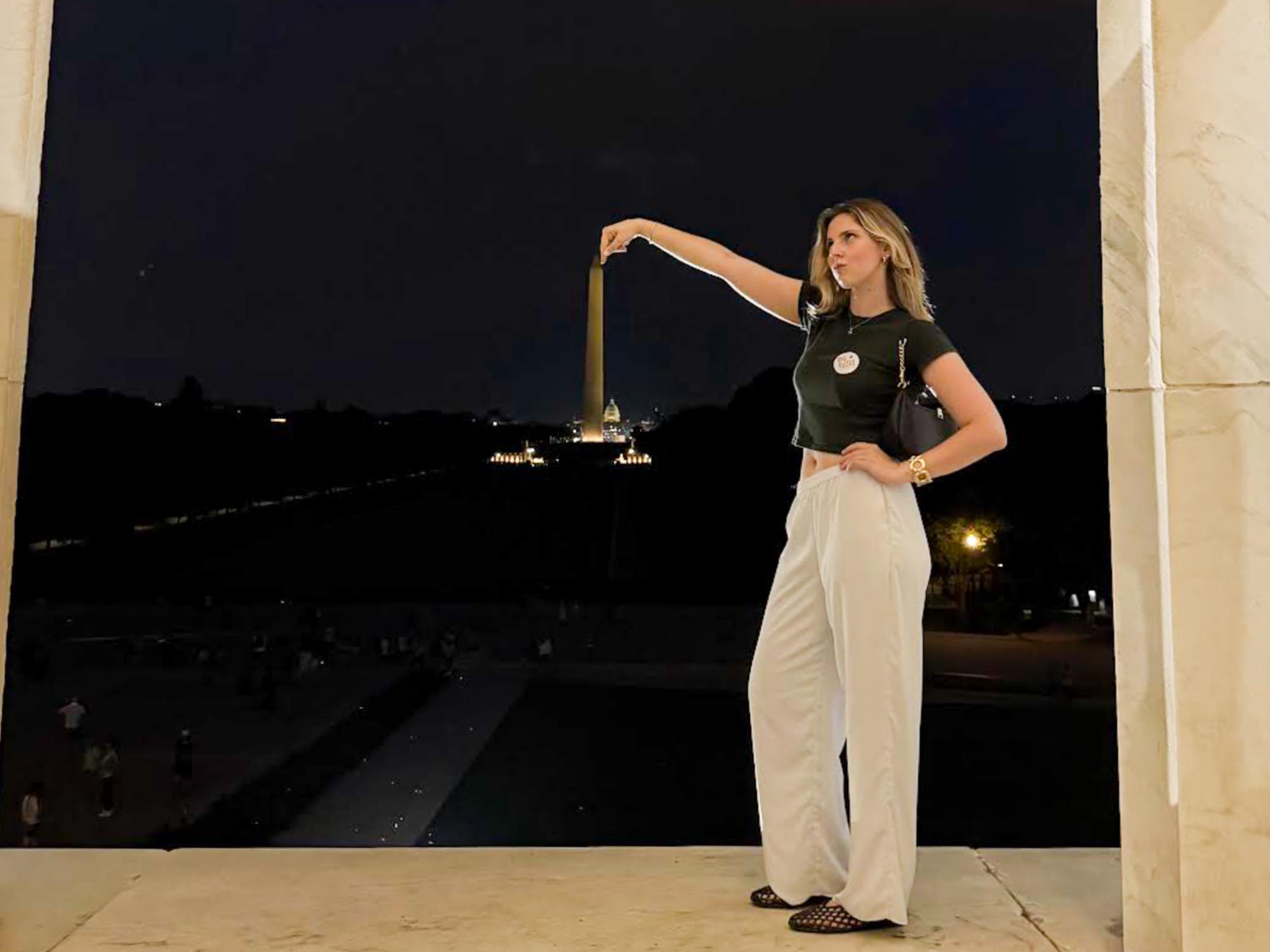 Reid-Pitta poses with hand up, with the Washington Monument in the distance. She wears white pants and a black top. 
