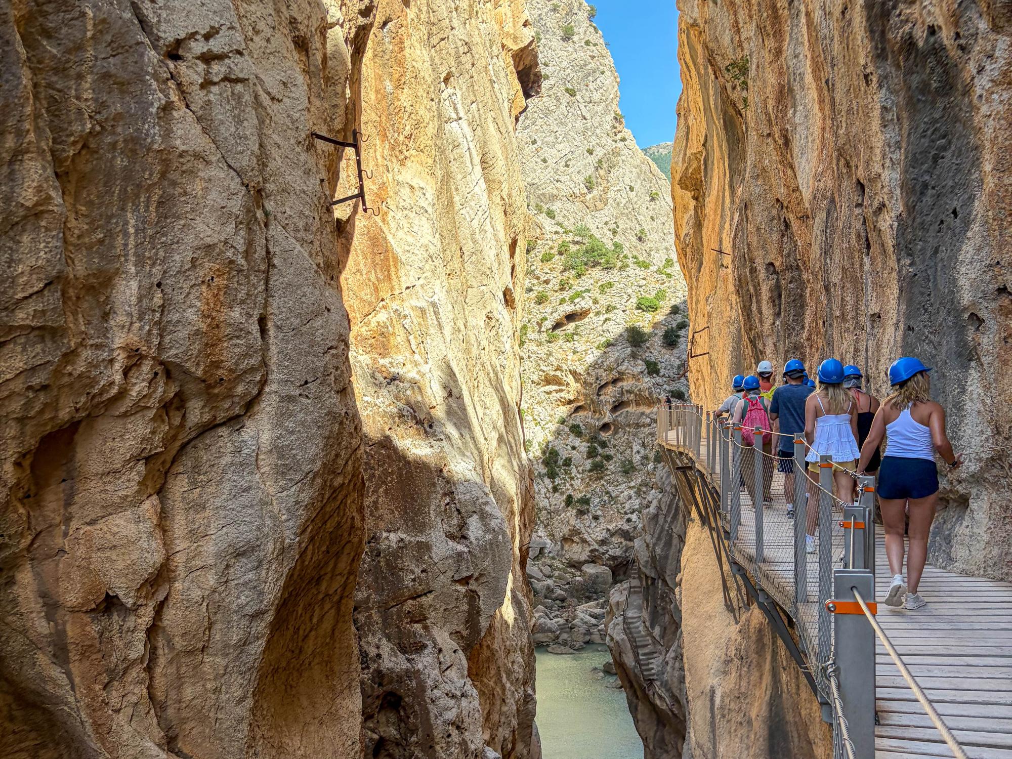 Tourists walk along a narrow, rocky bridge high above a rugged, canyon landscape.