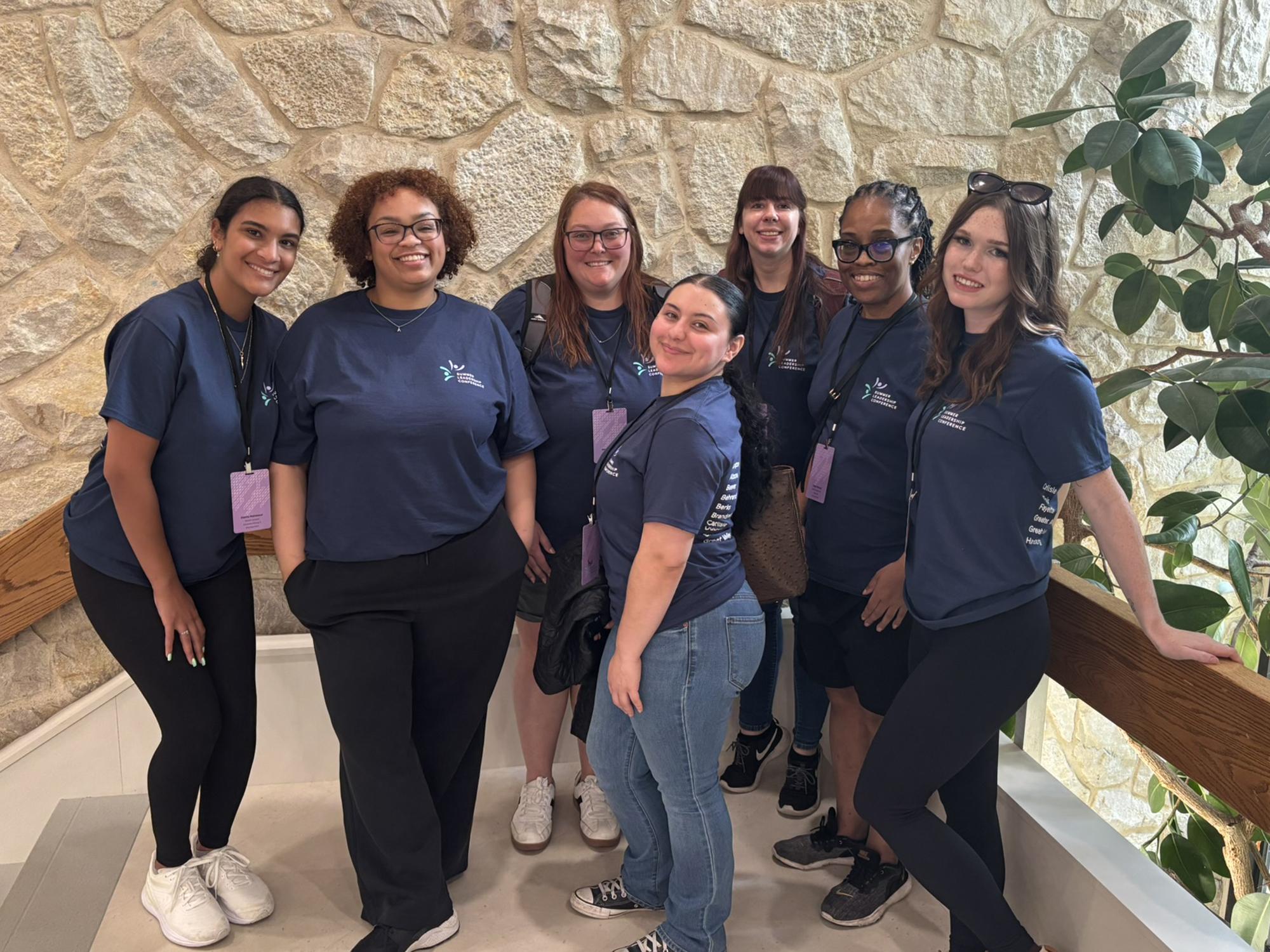 Seven women pose in front of a stone wall and plants.