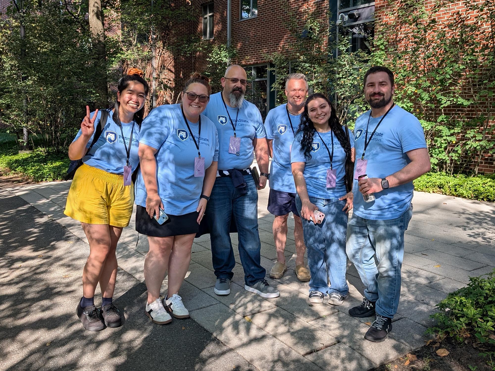 Six people wearing matching blue T-shirts pose for a photo in front of a brick building.