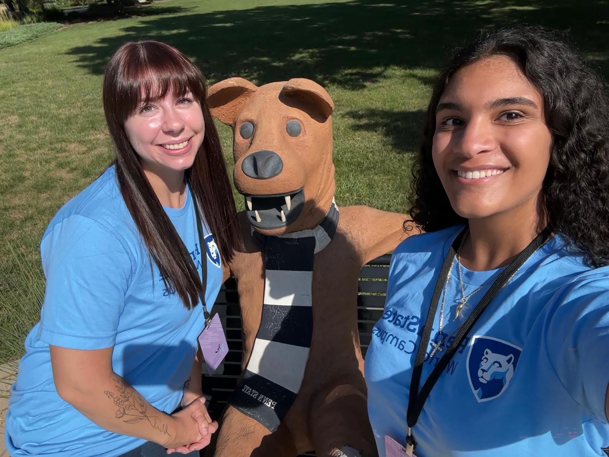 Two women sit on a bench with a statue of the Nittany Lion mascot.