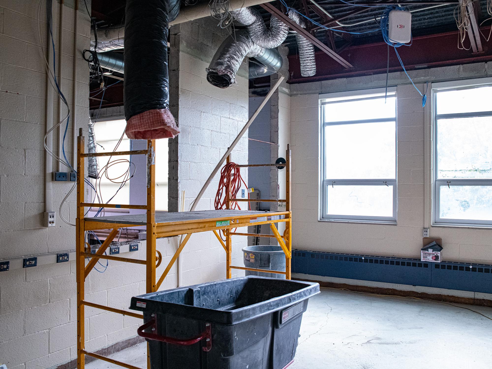 Interior room under renovation with exposed ceiling infrastructure, ductwork, electrical wiring, scaffolding, and construction equipment.