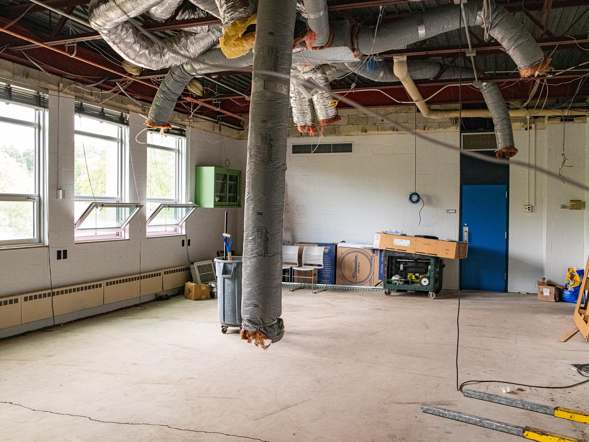 A former classroom or laboratory space undergoing renovation, featuring exposed ductwork, suspended utilities, construction materials, and open floor space.