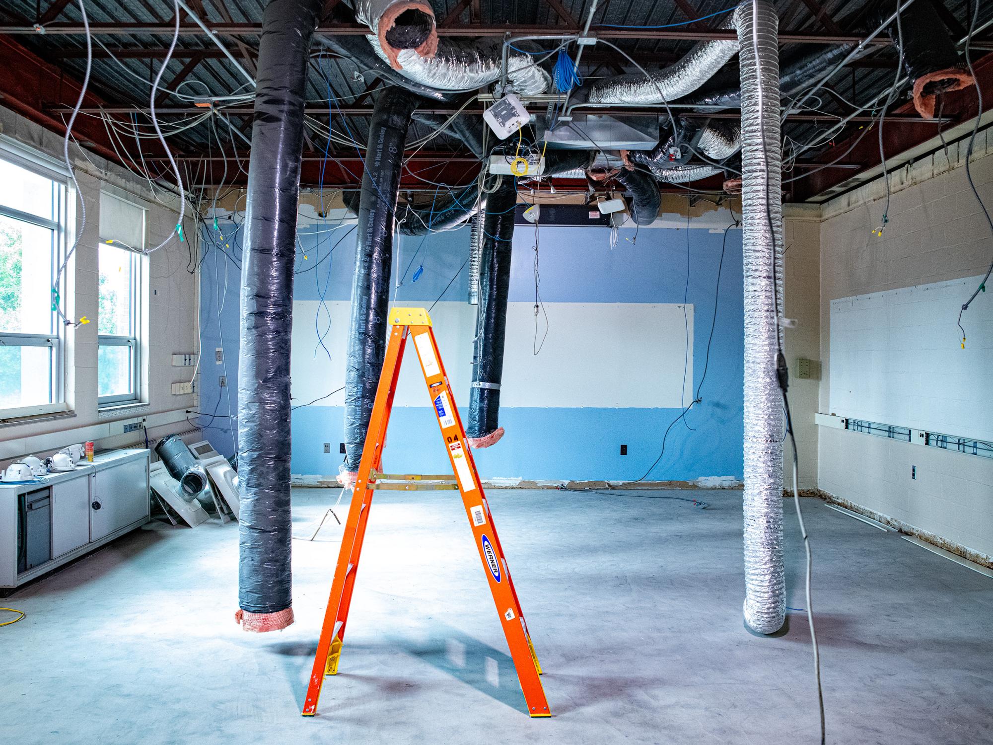 Academic laboratory space under renovation with exposed ductwork, wiring, ceiling infrastructure, and an orange ladder positioned in the center of the room.