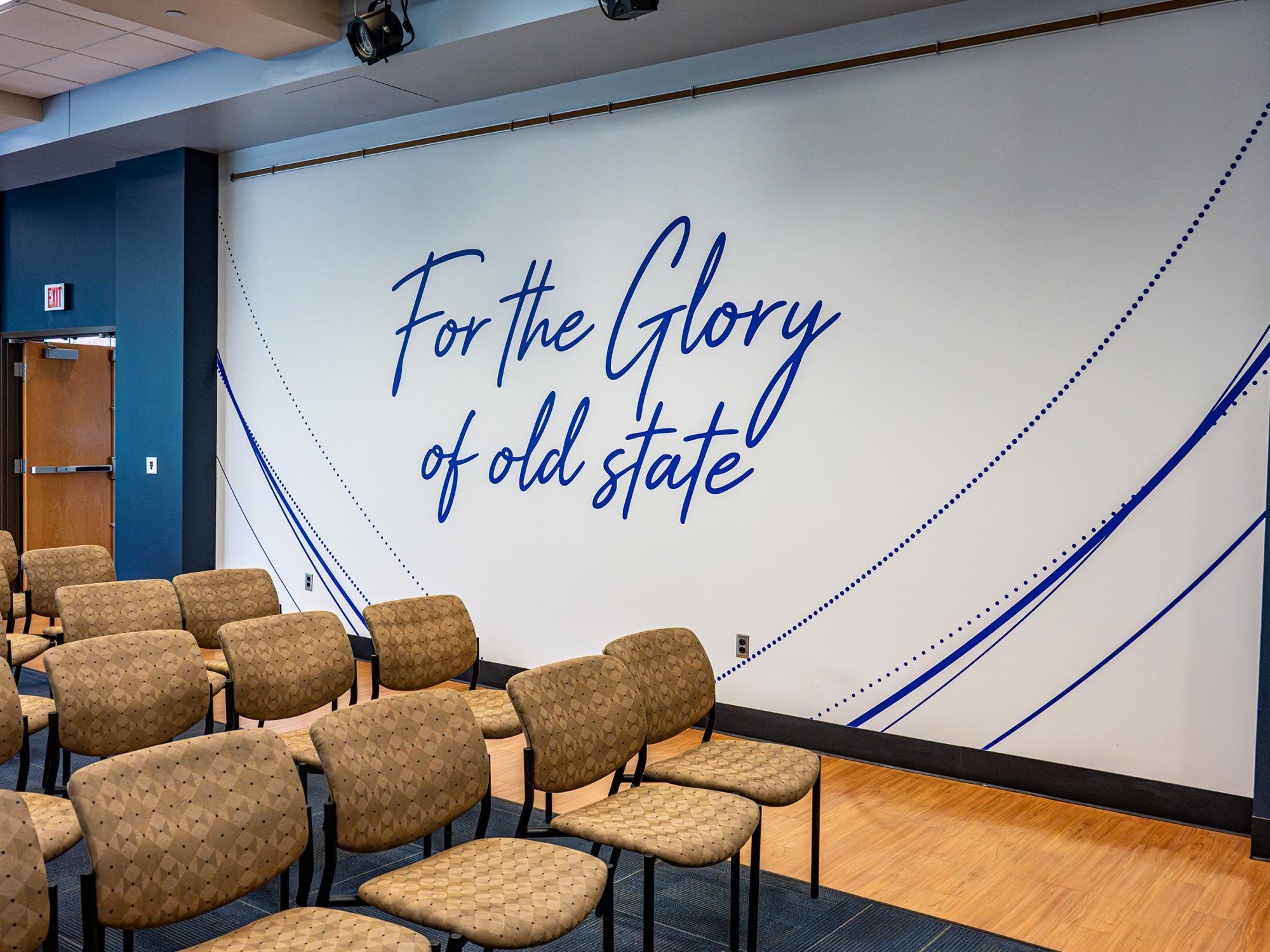 Rows of chairs face a newly renovated event space featuring a large wall graphic that reads “For the Glory of old state” in blue script.