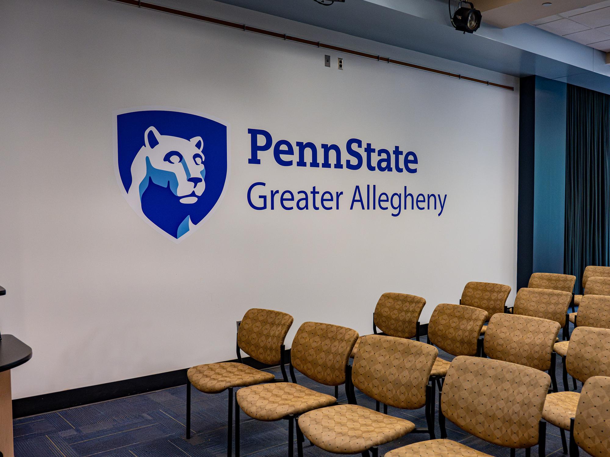 Penn State Greater Allegheny wordmark and Nittany Lion shield logo displayed on a white wall behind audience seating in the Student Community Center.