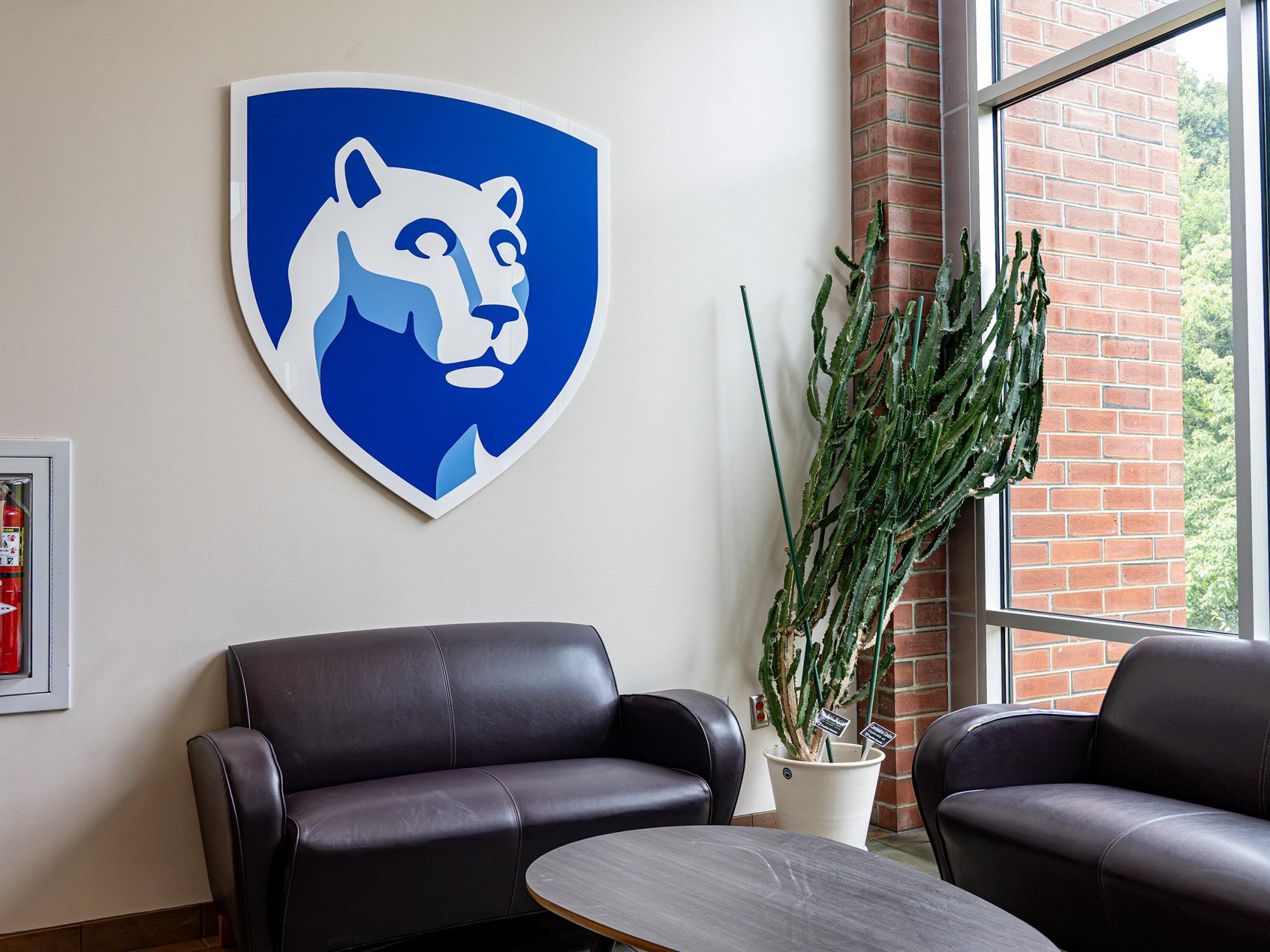 A seating area in the Student Community Center features a large Penn State Nittany Lion shield mounted on the wall above leather chairs and a coffee table near floor-to-ceiling windows.