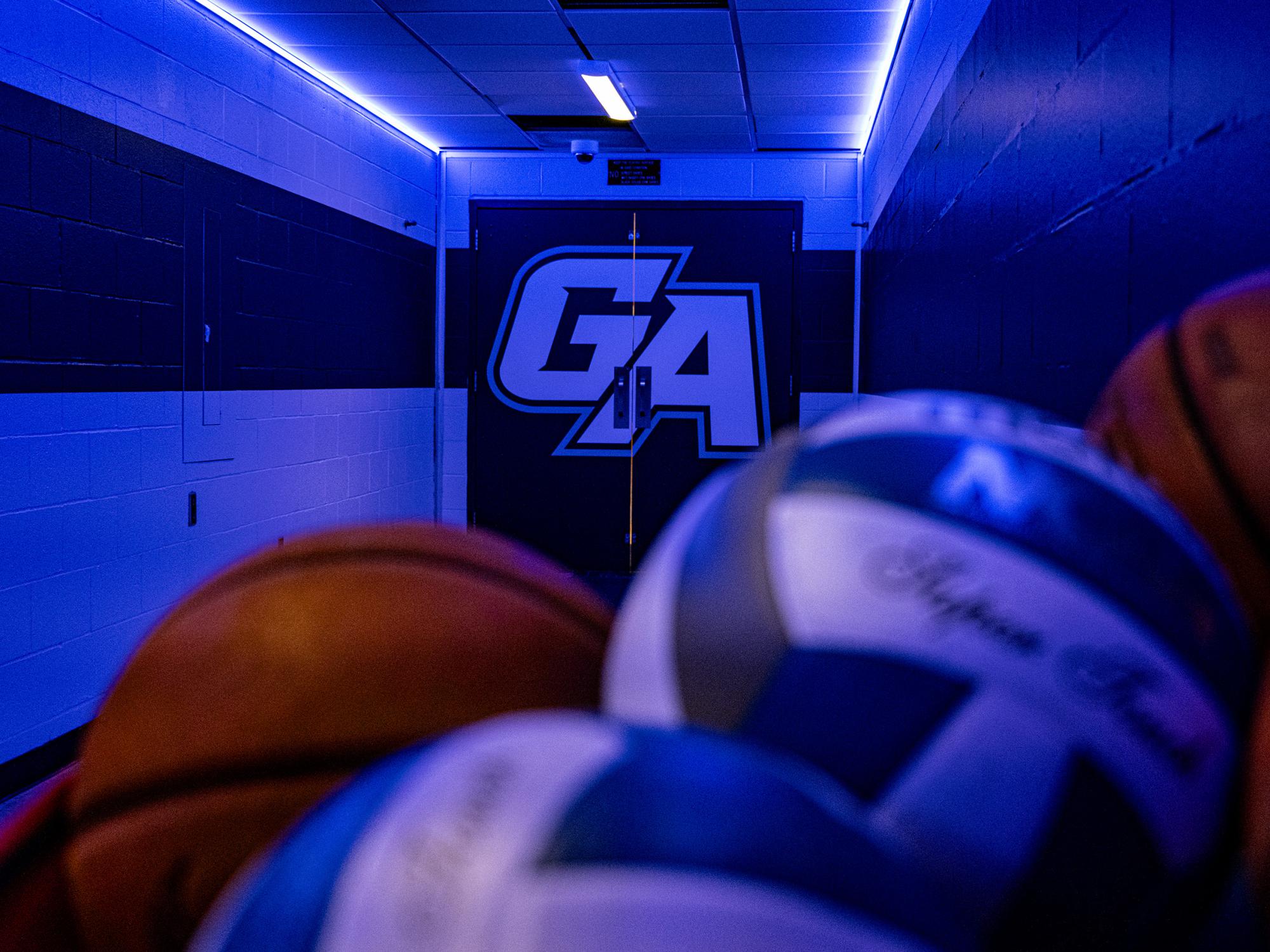 A tunnel in Wunderley Gymnasium features blue LED accent lighting and a large illuminated "GA" logo on the doors at the end of the hallway, with basketballs and volleyballs in the foreground.
