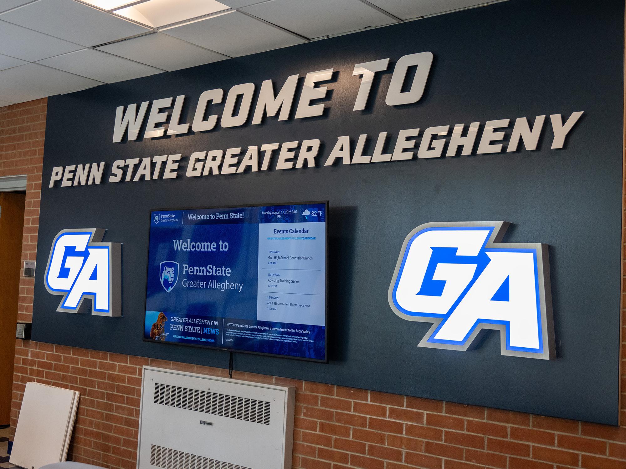A wall display in the Wunderley Gymnasium lobby features illuminated "GA" logos, a digital information screen, and large lettering reading "Welcome to Penn State Greater Allegheny."