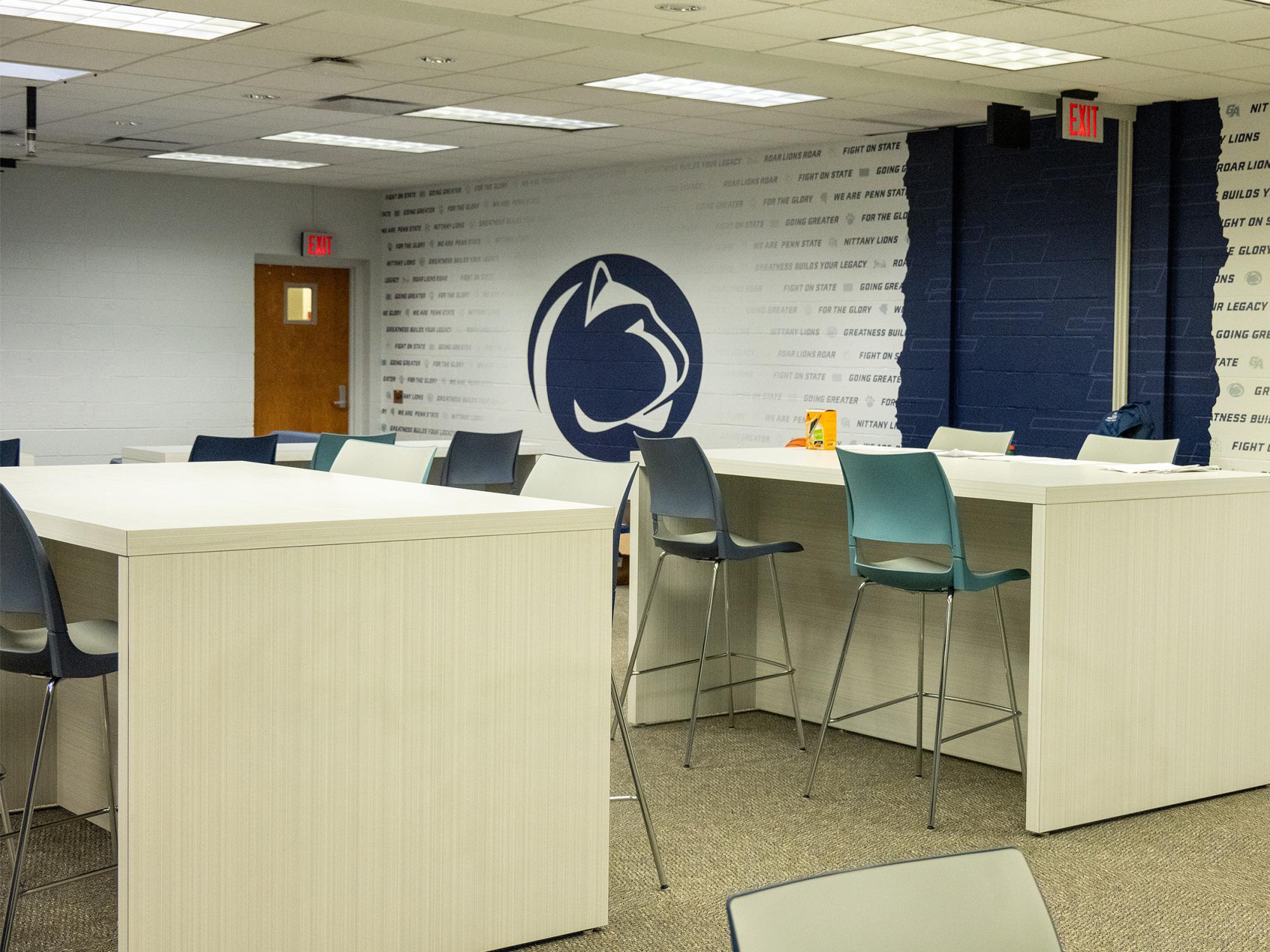 The renovated athlete lounge in Wunderley Gymnasium features new high-top tables, chairs, and Penn State-themed wall graphics, including a large Nittany Lion logo.