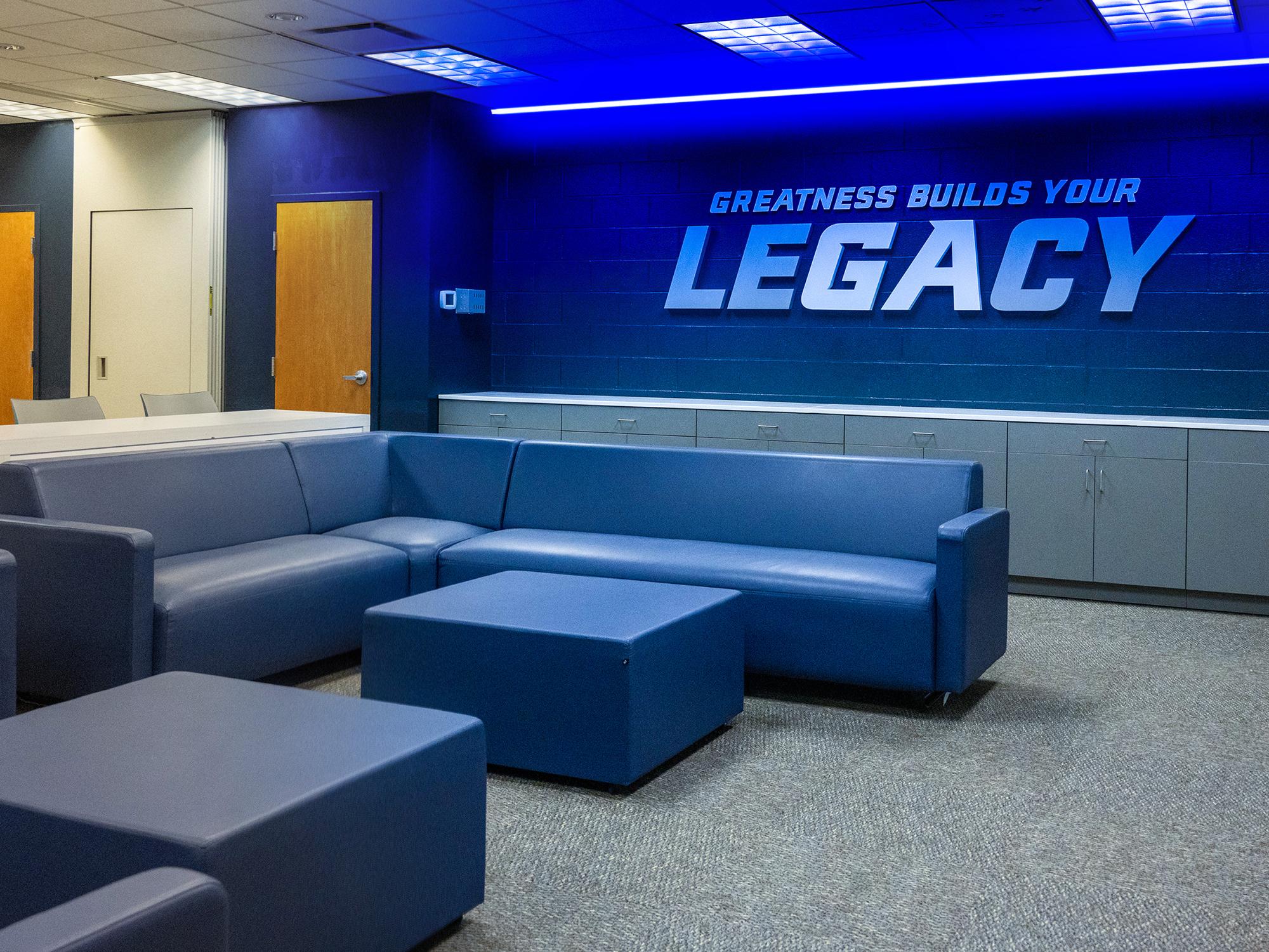 A renovated lounge space in Wunderley Gymnasium includes new seating, cabinetry, blue LED accent lighting, and a large wall graphic reading “Greatness Builds Your Legacy.”