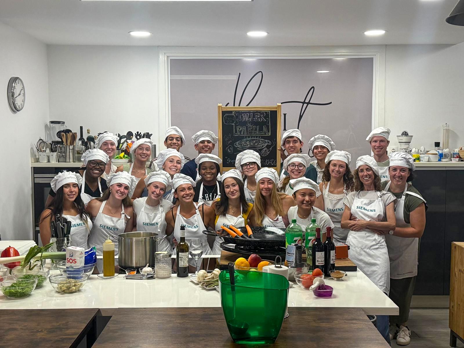 A group of people wearing white chef hats and aprons gather around a cooking station with ingredients, posing in a kitchen.