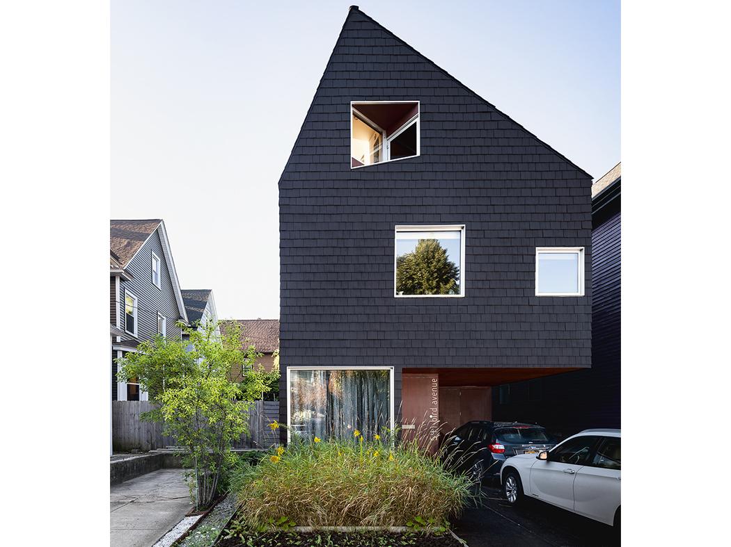 Exterior of a modern three-story house with dark black shingles, an asymmetrical pitched roof, square windows, and a cantilevered driveway with parked cars.
