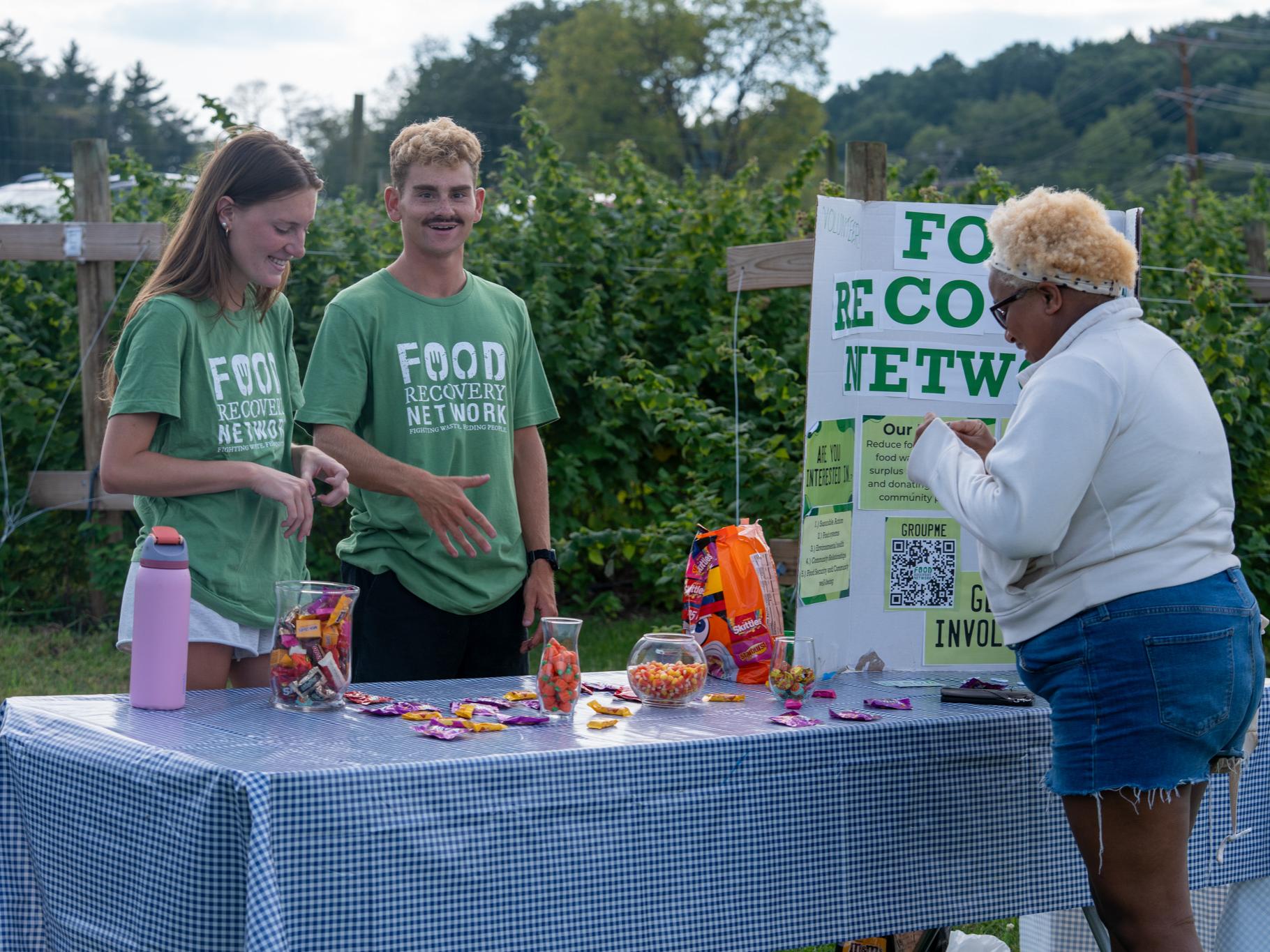 Two Penn State Food Recovery Network members wearing green T-shirts stand behind an information table at Harvest Fest at the Dr. Keiko Miwa Ross Student Farm. The table is covered with a blue-and-white checkered cloth and displays jars and bags of candy alongside a Food Recovery Network poster as they speak with an attendee.
