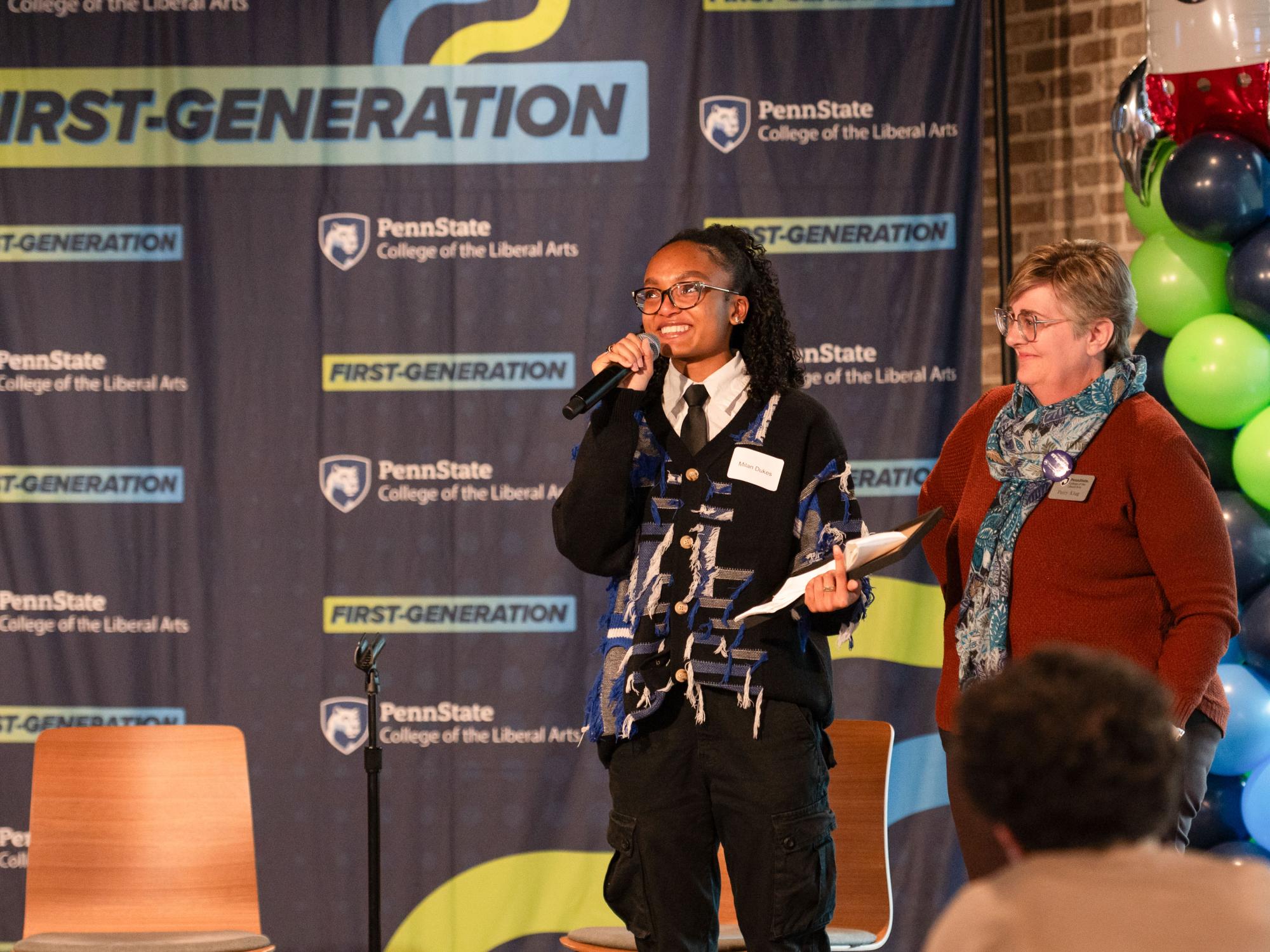 Milan Dukes, center, smiles while holding a microphone next to Patty Klug, director of the Liberal Arts Chaiken Center. Balloons and a banner repeating “First-Generation” and “Penn State College of the Liberal Arts” are visible behind them.