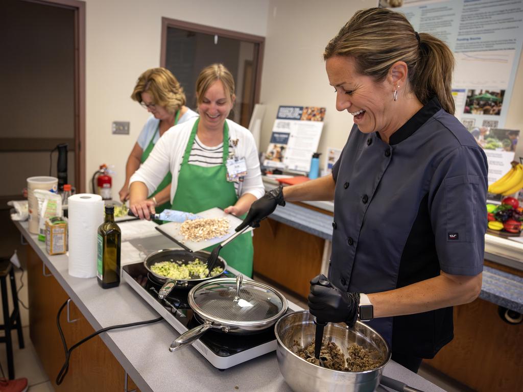 Hillary Amici, right, an education program specialist at Penn State College of Medicine, laughs as she stirs two pans on a stovetop at the same time. Two women wearing aprons cook next to her.