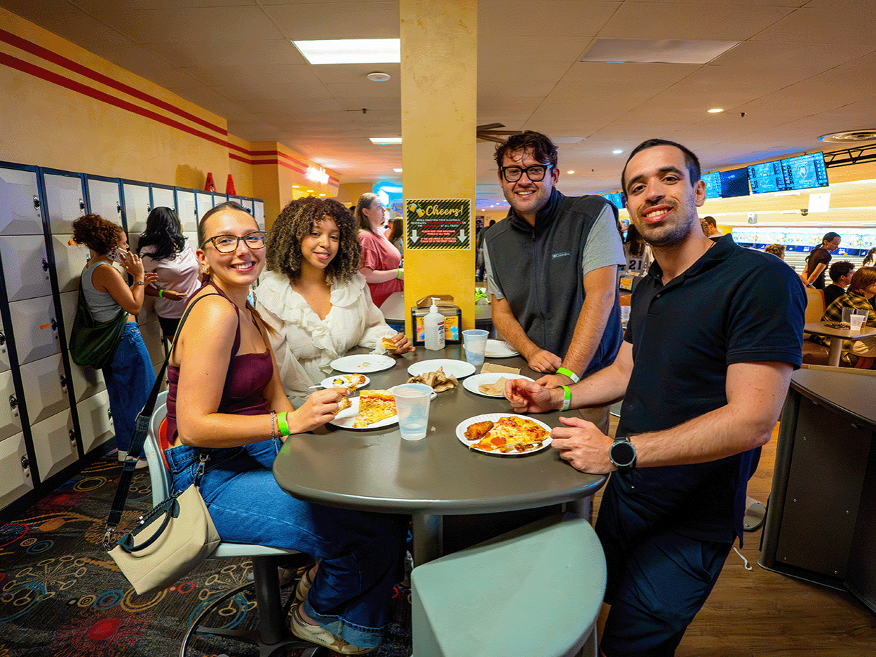 Students eating pizza and chicken wings around a table at a bowling alley