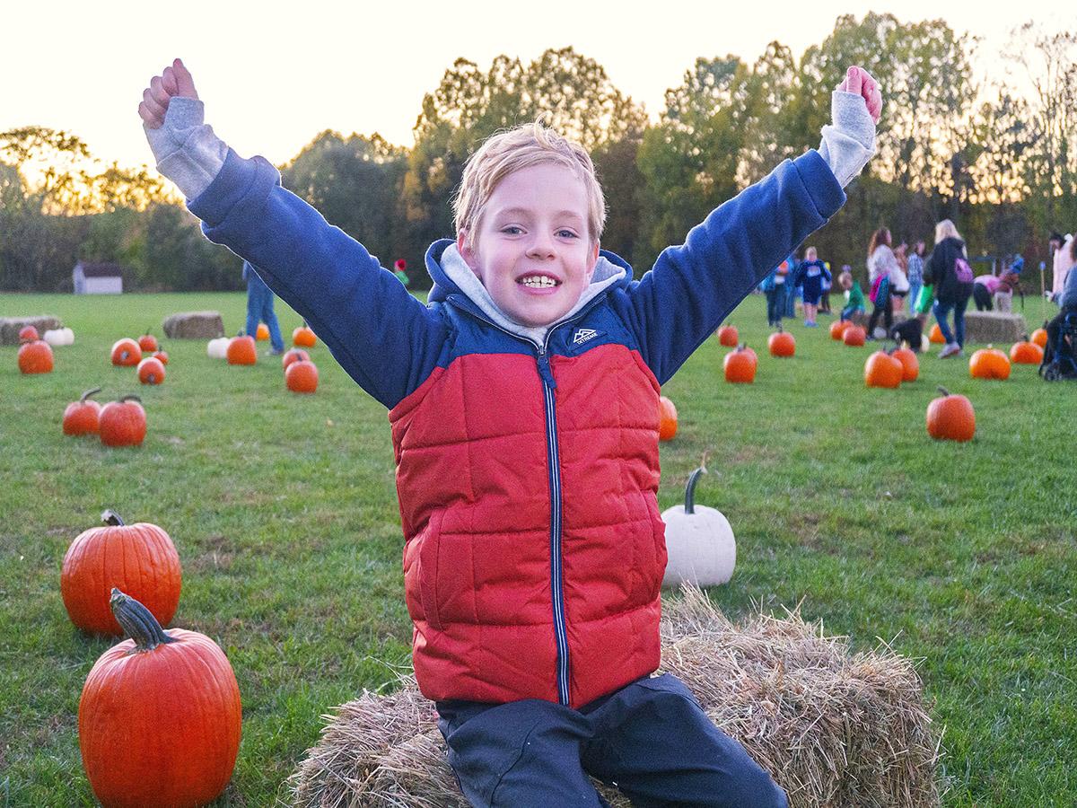 Child sits on hay bale in the pumpkin patch with arms extended and smiling