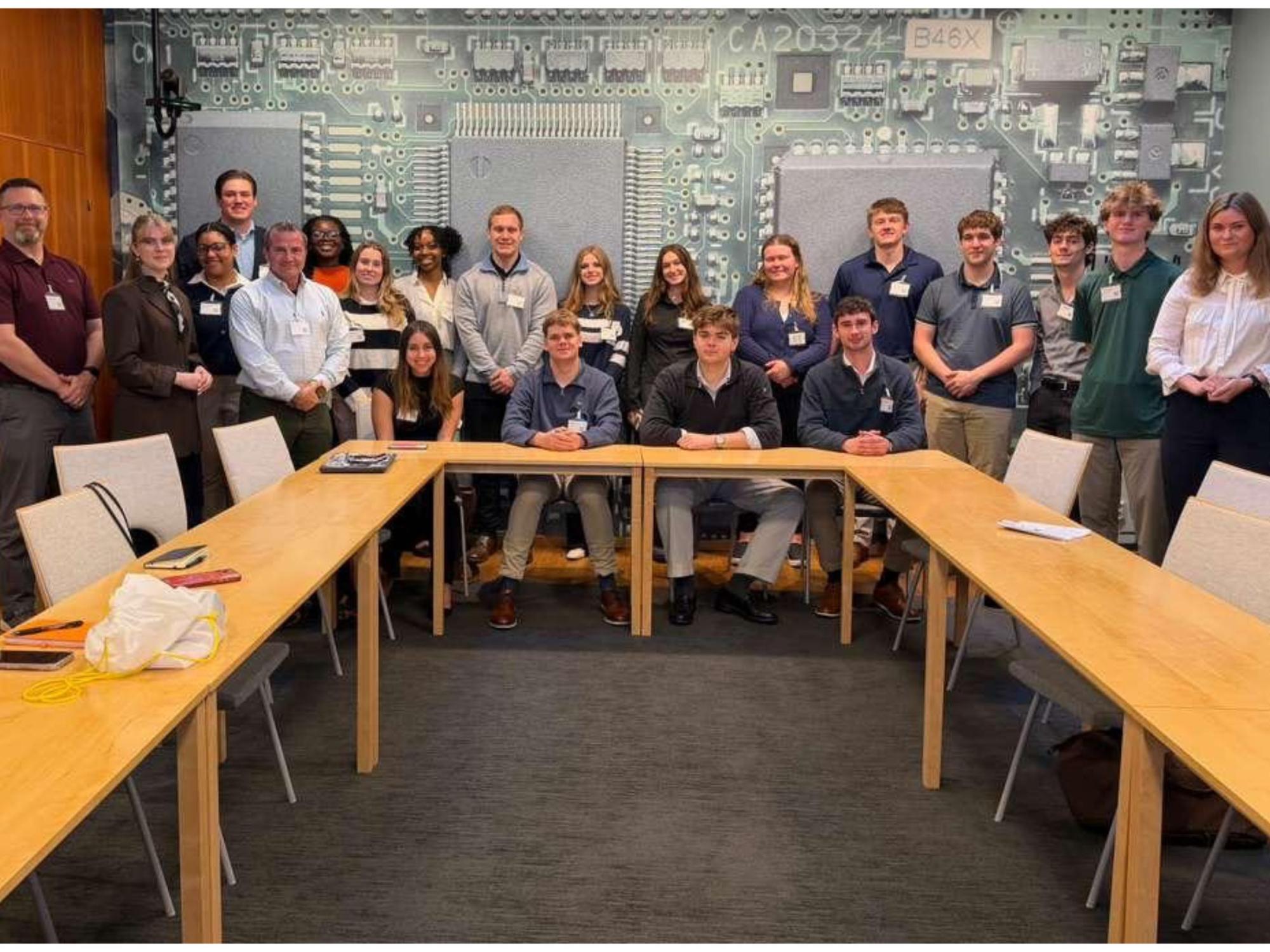 a large group seated around a conference table with a printed circuit board all behind them