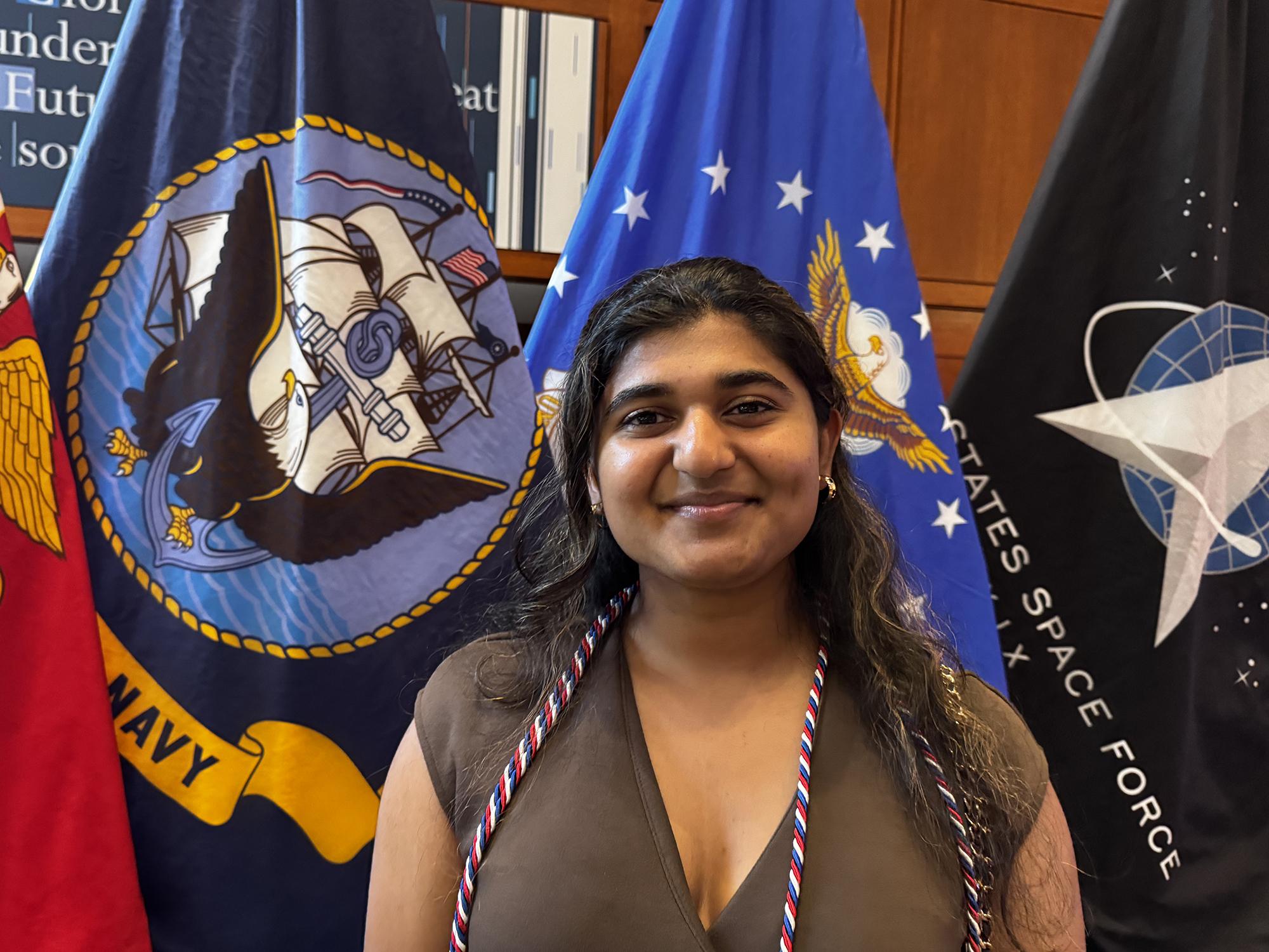 A woman wearing red, white, and blue honor cords poses in front of a group of flags.